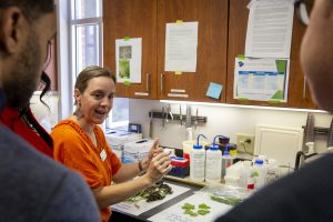 Carrie Harmon in a lab setting speaks to a group of students gathered around her, with laboratory equipment and supplies visible in the background.