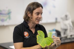Carrie Harmon wearing a black collared shirt holds a large green leaf while speaking in a classroom or lab setting, with a whiteboard and equipment visible in the background.