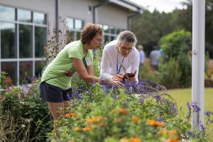 Two women side by side looking at a green bush with purple flowers.