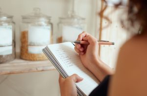 budget planning, making shopping list and managing household expenses to save money. Financial accountability at home. Woman making shopping list for groceries on a notebook to plan a meal for dinner.