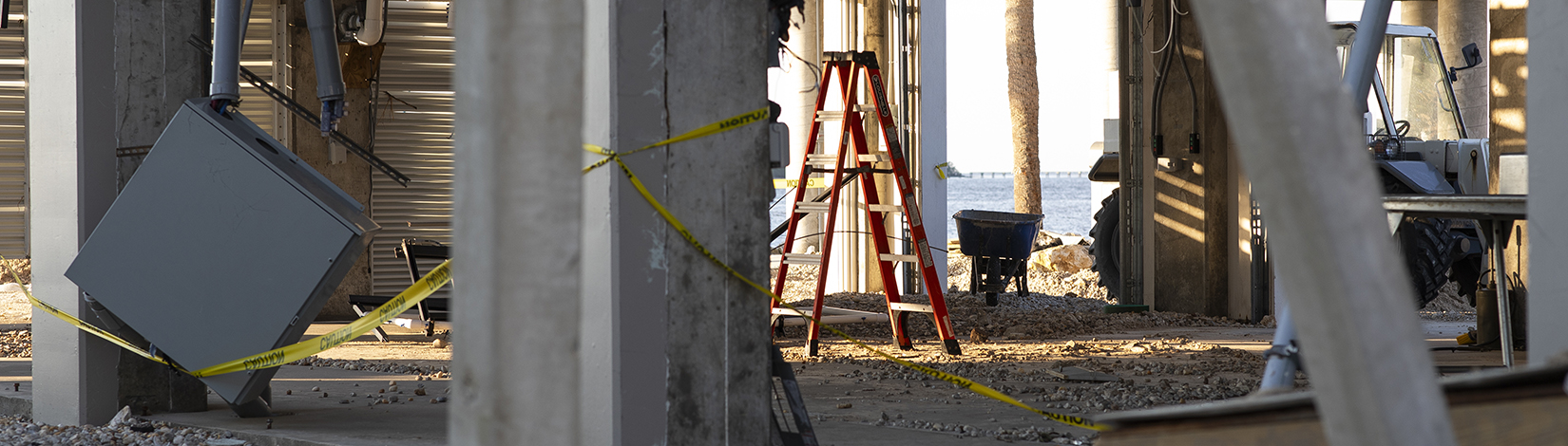 hurricane damage beneath a raised coastal structure with concrete pillars, a red ladder, caution tape, and a damaged utility box amid gravel and debris.