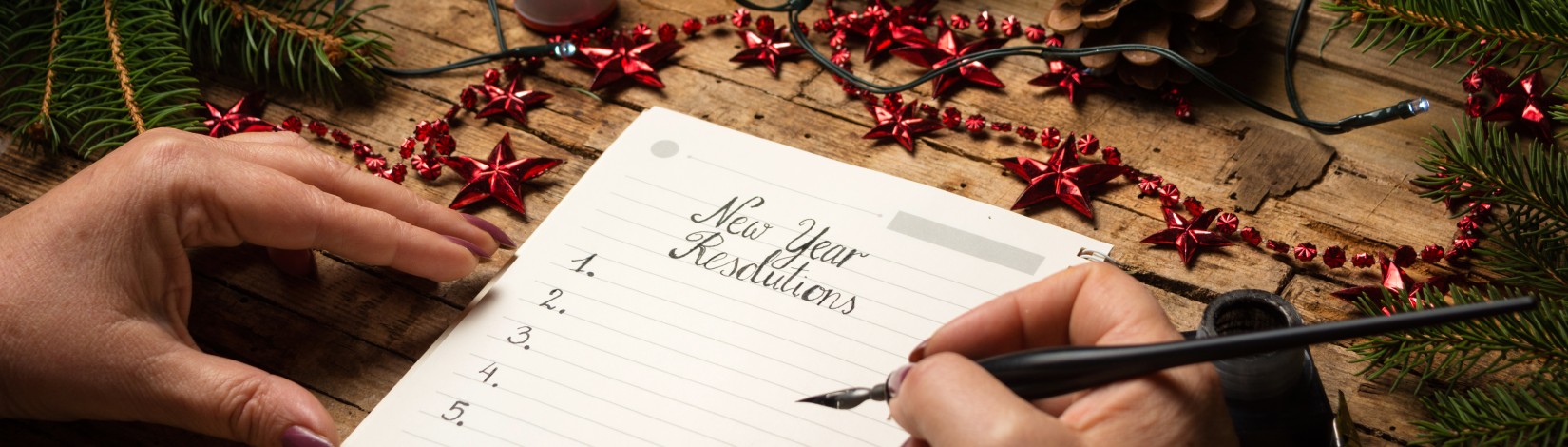 Hands writing a “New Year Resolutions” list in a notebook on a rustic wooden table, surrounded by pine branches, pinecones, wrapped gifts, red star garlands, and holiday lights. Photo by Victor Moussa. AdobeStock