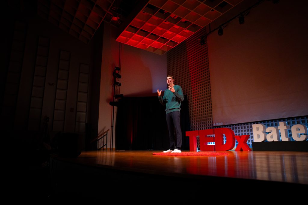 A man stands on stage. The stage has a decoration that reads "TEDx Bates."