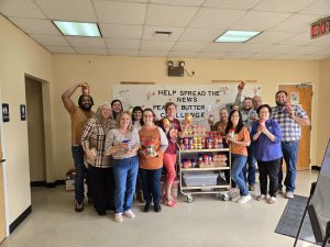 Group of people smiling and holding peanut butter jars on both sides of a table filled with peanut butter jars in an indoor setting, with a banner behind that reads “Help Spread the News: Peanut Butter Challenge.