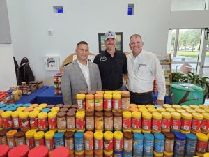 Three men smiling behind a row of multicolored peanut butter jars in an indoor setting.