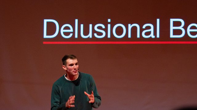 A man stands in front of a presentation screen. The screen displays the words "delusional belief."