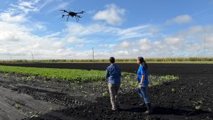 Two people stand in a cultivated farm field, watching a large drone hover above the crops under a bright blue sky with scattered clouds.