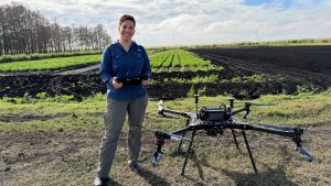A person stands in a cultivated field holding a drone controller, with a large multi-rotor agricultural drone resting on the ground beside them; neat rows of green crops stretch into the background under a partly cloudy sky.