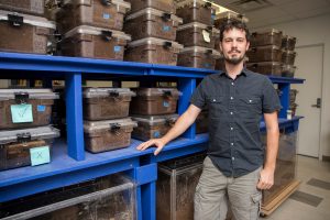 A man stands next to blue shelves filled with clear plastic containers. He wears a black shirt and beige shorts. The indoor setting is a research lab.