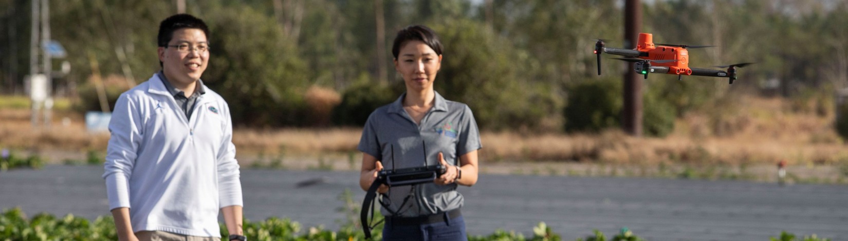 Two people standing in an open field while flying a drone; the woman in the foreground is holding the controller as the drone hovers in front of them
