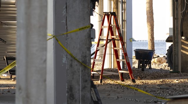 hurricane damage beneath a raised coastal structure with concrete pillars, a red ladder, caution tape, and a damaged utility box amid gravel and debris.