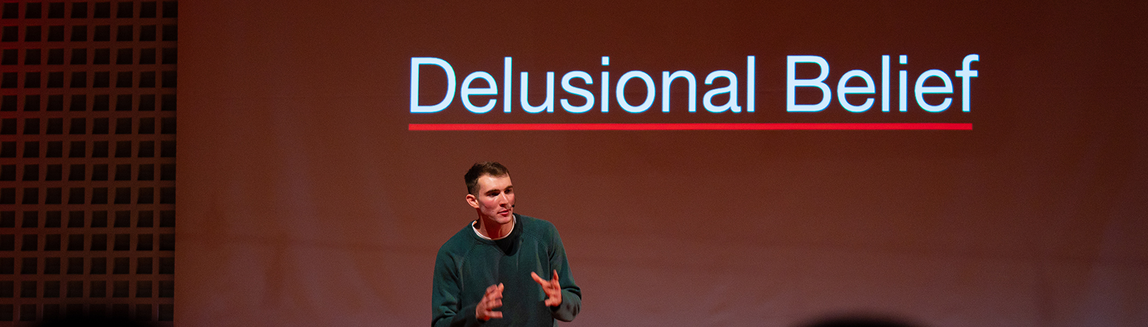 A man stands in front of a presentation screen. The screen displays the words "delusional belief."