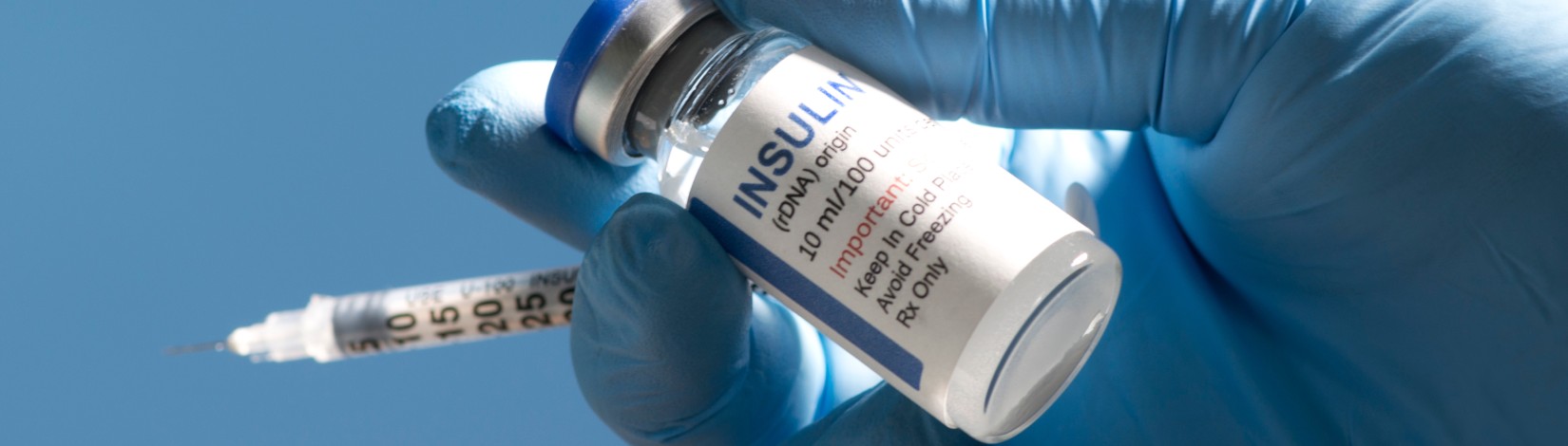 A gloved hand holds a vial of insulin and a syringe against a clear blue background. The vial label shows dosage and storage instructions.