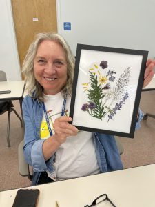 Person smiling holding their picture frame. Their art has various pressed floral material of flowers and foliaged arranged in a decorate design.