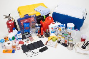 photo of a hurricane kit containing a cooler, gas can, first aid supplies, water, canned goods, fire extinguisher, TP, paper products, chargers and more.