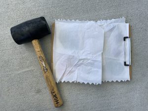 2 sheets of paper towel laying on top of paper with flowers taped to it. Sitting next to it is a rubber mallet.
