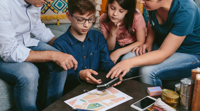 a man wearing a blue shirt and jeans and a woman wearing a blue shirt and jeans are showing a boy with brown hair and glasses how to use a weather radio. There is also a girl with brown hair and a red shirt in photo along with various hurricane supplies
