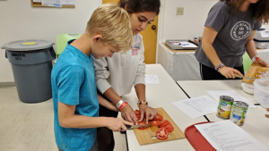 A teen 4-H member teaching a younger 4-H member how to cut tomatoes. 