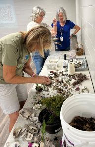 three women standing around a table full of plants and decorations to be used for building the plant vogs