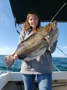 a women on a boat holding up a large gag grouper. It is dark tan with black markings 