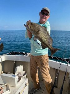 Man holding up a Gag Grouper on a boat and smiling. The fish is about 20 inches in length and dark brown in color. 