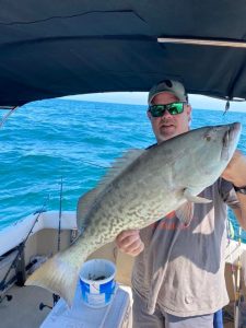 Man holding up a Gag Grouper on a boat. The fish is about 30 inches in length and tan to grey in color. 