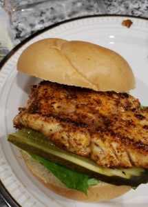 Cooked blackened fish fillet on a hamburger bun with pickle and lettuce sitting on a plate 