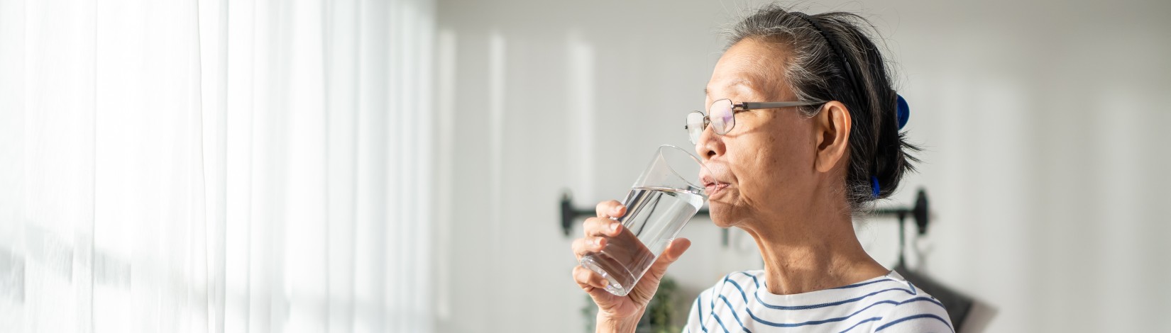 Asian senior mature woman drinking a glass of water in kitchen at home.