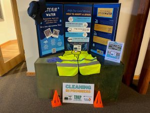 Trifold display promoting the Adopt-A-Drain program, showcasing materials such as sunhat, cones, safety vest, gloves, "Cleaning in Progress" sign, and educational information about stormwater and how to get involved with the Adopt-A-Drain program on the trifold display.