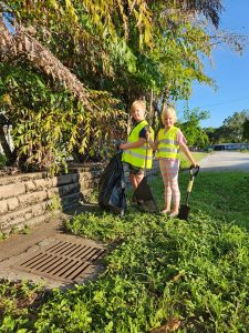 A young boy and girl standing with the boy holding a large black garbage bag and girl holding a shovel, wearing neon yellow safety vests standing in front of clean storm drain they just cleared.