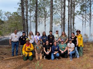 Group of male and female adults standing out in the woods in front of a prescribed fire.
