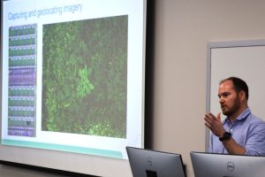 photo of Dr. Ian Small speaking at a lectern on the right side of the image, with a projector screen on the left side