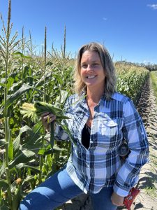 female standing in a sweet corn field holding an ear of corn