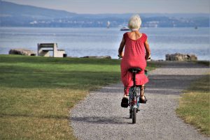 a woman in a red dress rides a bicycle along a path by a lake. [credit: pixabay.com, julita]