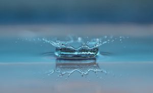 a drop of water splashes into an otherwise smooth pool of blue water. [credit: pixabay.com, roegger]