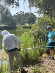 Volunteer Anne Maloney stands at the edge of a pond next to the vegetation with a student wearing a hoodie and pants as he holds a long handled dip net.
