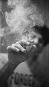 closeup of a cigarette in a hand amidst a haze of smoke, with the smoking man in background. [credit: unsplash.com, arun anoop]