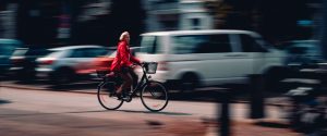 a woman wearing a red jacket rides a bike briskly past parked cars in an urban setting. [credit: unsplash.com, kieran sheehan]