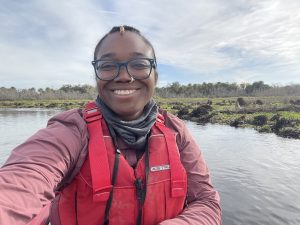 A photo of a woman with glasses smiling at the camera, with a life vest, in a kayak on the water.