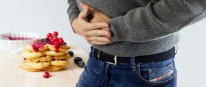 a man holds his aching stomach, with food on a table in the background. [credit: pxhere.com]