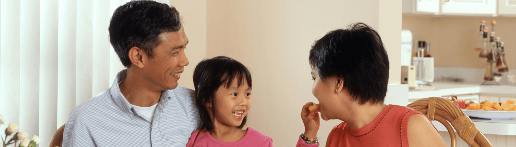 a young girl sits at a glass-top table with a man and woman, smiling as she hand-feeds the woman. [credit: unsplash, national cancer institute]
