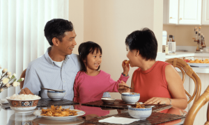 a young girl sits at a glass-top table with a man and woman, smiling as she hand-feeds the woman. [credit: unsplash, national cancer institute]