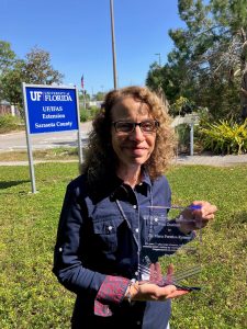 Maria Portelos-Rometo holding retirement plaque in front of UF/IFAS Extension Sarasota County sign. [credit: uf/ifas extension sarasota county]