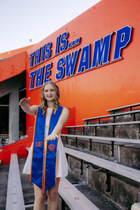 Hailey stands in the University of Florida football stadium and does a gator chomp. She hold her right arm above her left in an outstretched manner to form an alligator mouth.