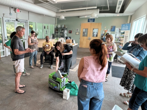 Eight people stand in a circle, watching a female staff member remove items from a green rolling cart and explain how to use them.