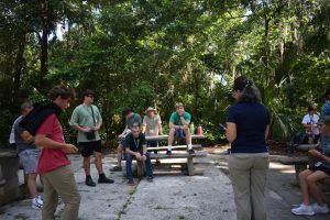 A group of kids circle around the presenter and sit on picnic tables.