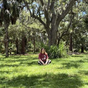 A woman sitting in a grassy field with large oak trees meditating.