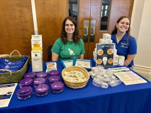 Two women smile behind a table with reusable snack containers, LED lightbulbs and nightlights, cooling towels, and printed information.