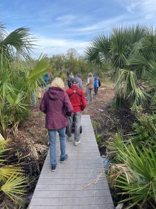 An image of a group walking across a bridge in a natural area, with a person in a wheelchair being pushed across the accessible bridge.
