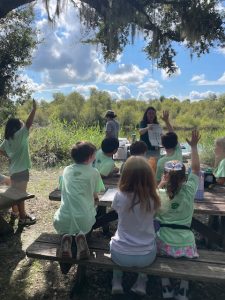 Students raise their hands to answer a question while sitting at a picnic table outdoors. 